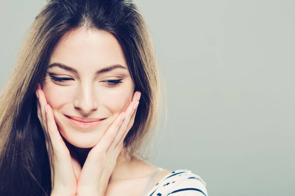 Eine junge Frau mit langen braunen Haaren berührt sanft mit beiden Händen ihre Wangen und lächelt sanft, während sie nach unten blickt. Sie strahlt das natürliche Leuchten aus, das von RINGANA Frischekosmetik inspiriert wurde, und steht vor einem hellgrauen Hintergrund.