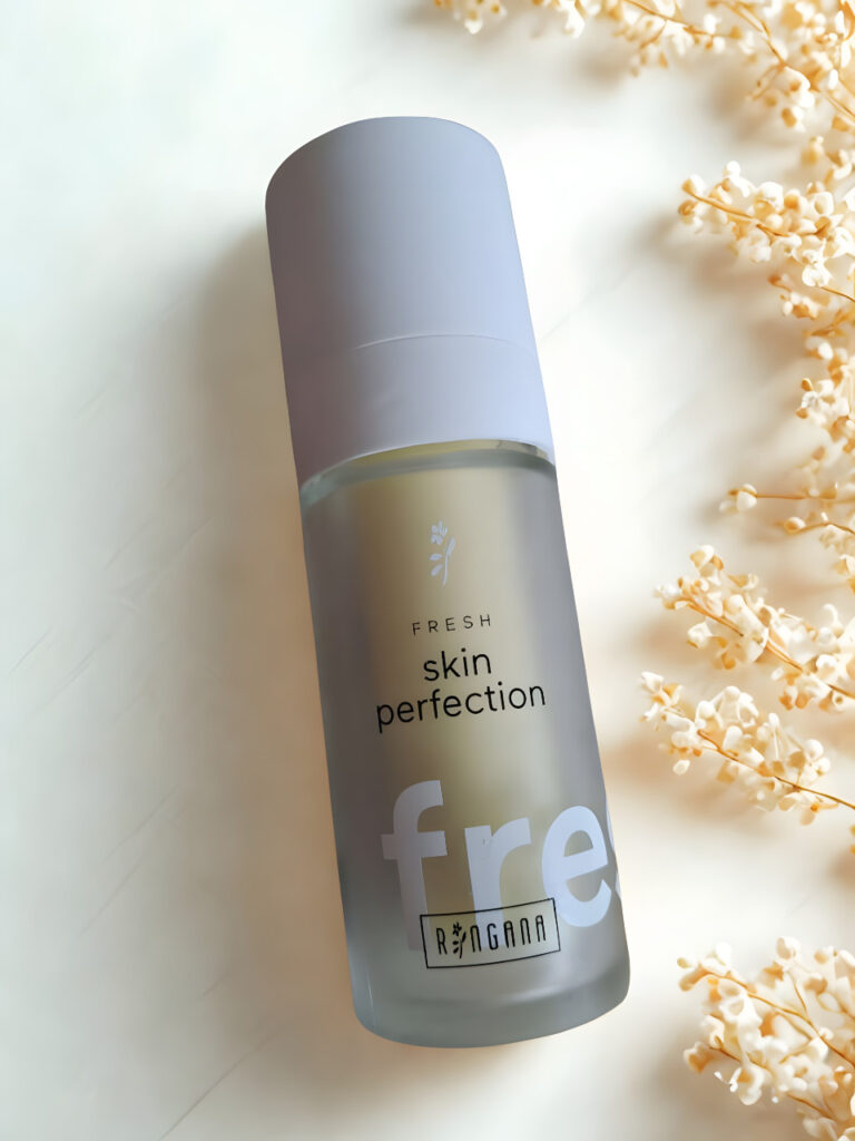 Ein Flakon des Serums Fresh Skin Perfection von Ringana steht auf einer hellen Fläche mit weichen Schatten und zarten, getrockneten weißen Blüten daneben - entdecken Sie mehr RINGANA Frischekosmetik im ringana online shop.