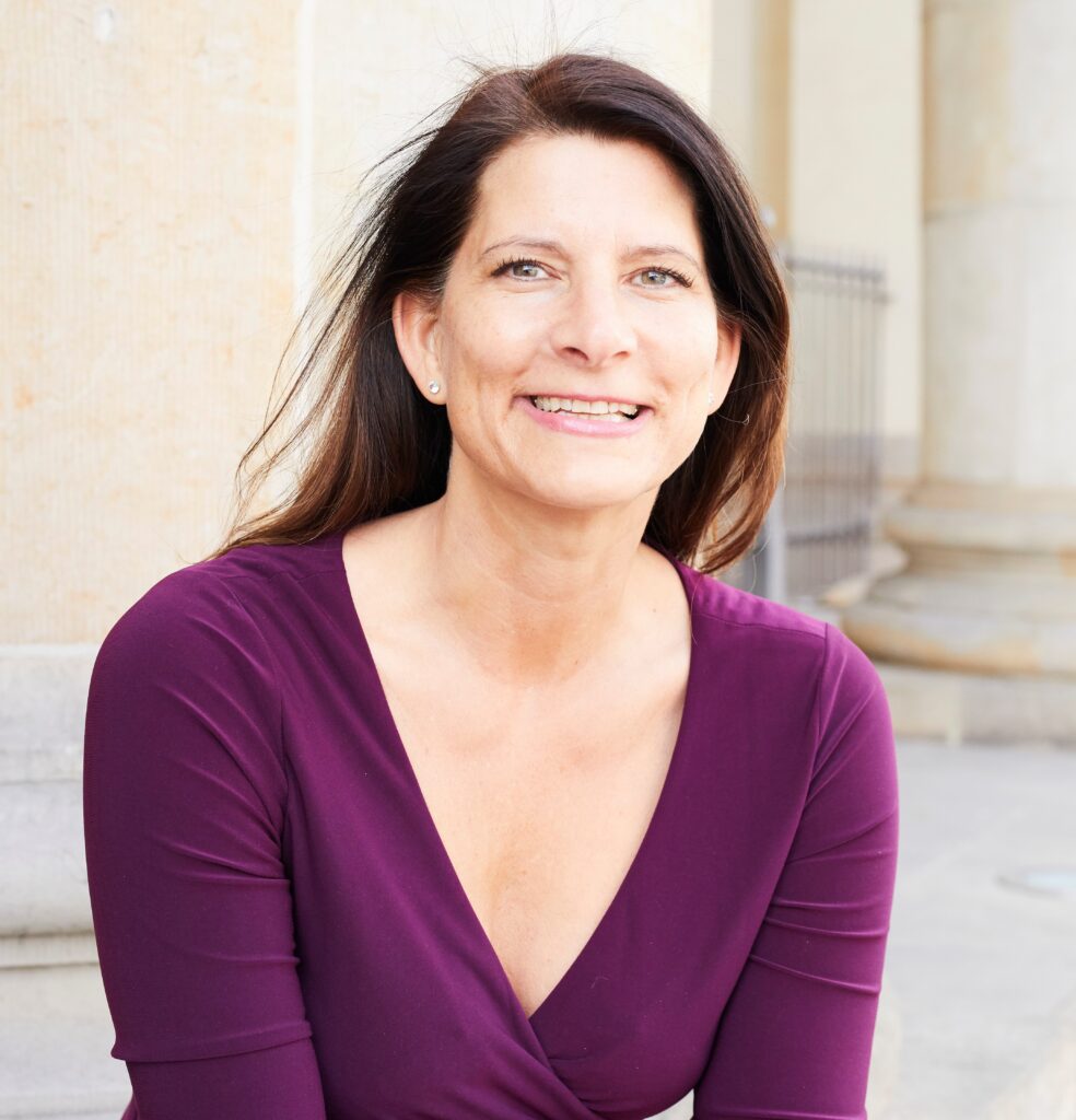 Stefanie Rensch, RINGANA Partnerin seit 2016, Network Marketing Profi und in der Zielstufe 8 von 10 mit V-Ausschnitt trägt, lächelt, während sie im Freien vor einem hellen Steingebäude sitzt. Sie strahlt eine Zuversicht aus, die man oft bei erfolgreichen Network-Marketing-Profis sieht.