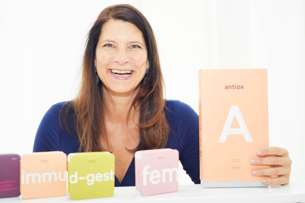 Eine lächelnde Frau mit langen braunen Haaren hält eine pfirsichfarbene RINGANA-Antiox A-Dose. Vor ihr stehen drei weitere bunte RINGANA-Dosen mit den Aufschriften immu-d-gest, fem und min.