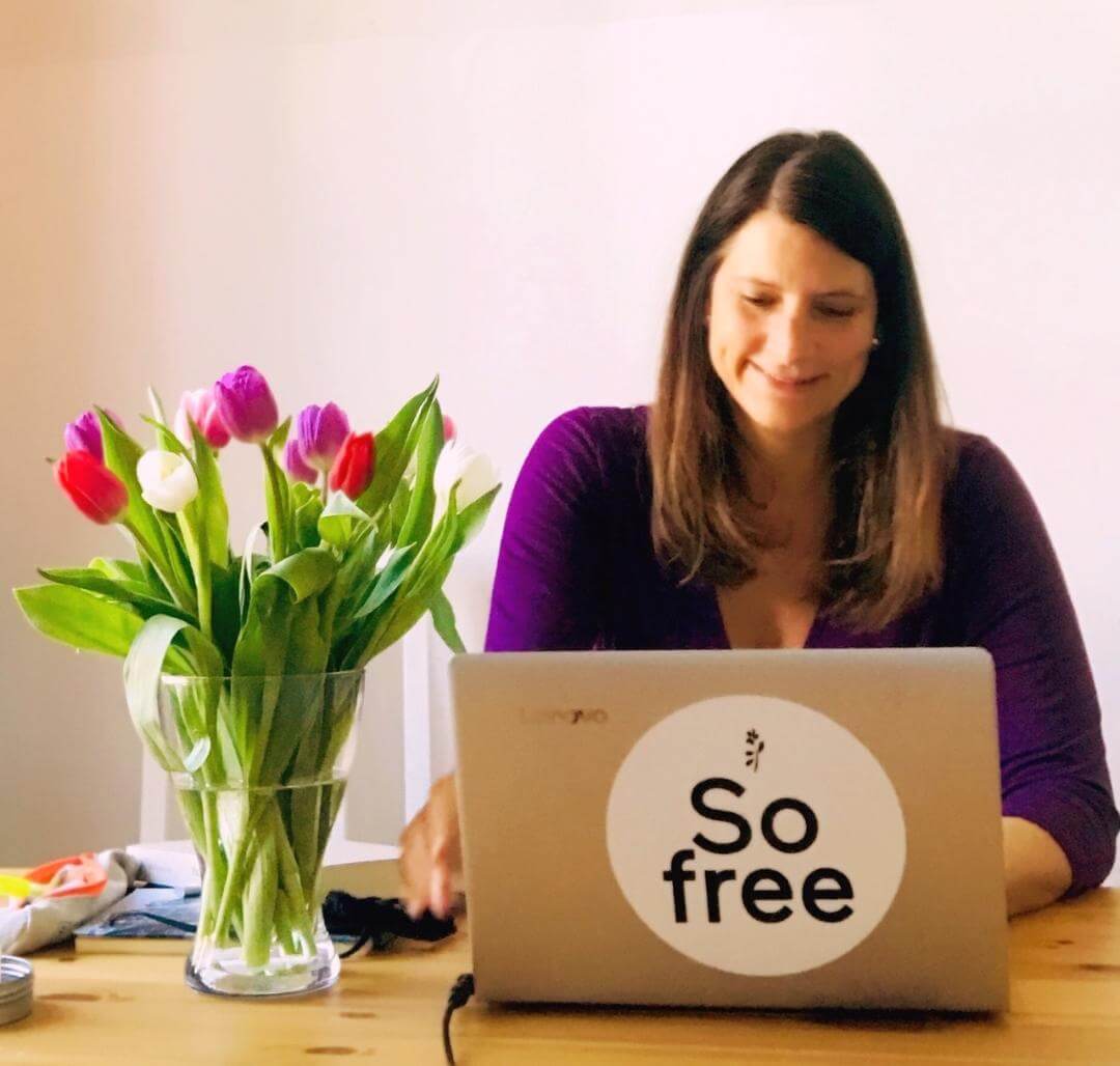 Eine Frau mit braunen Haaren sitzt an einem Holztisch und benutzt einen Laptop mit einem großen So free-Aufkleber und einem RINGANA-Notizbuch neben sich. Eine Glasvase mit bunten Tulpen steht auf dem Tisch. Der Hintergrund ist schlicht und hell.