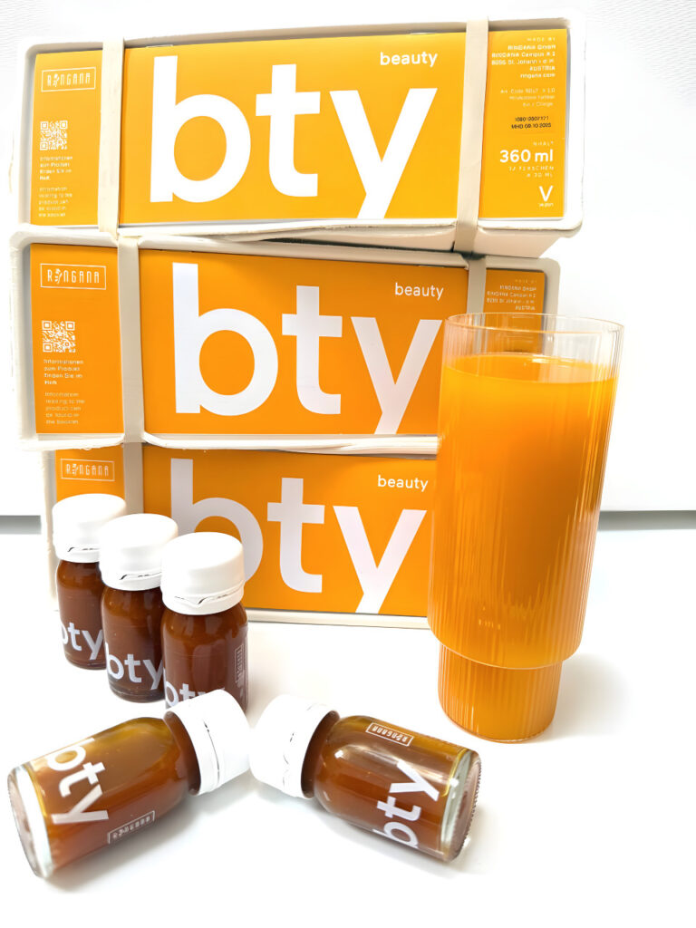 Im Hintergrund stapeln sich drei orangefarbene und weiße bty-veganes Kollagen Boxen. Davor liegen fünf kleine Flaschen mit der Aufschrift bty-Kollagen vegan Nahrungsergänzung verstreut neben einem hohen Glas mit orangefarbener Flüssigkeit. Die Szene ist hell beleuchtet.