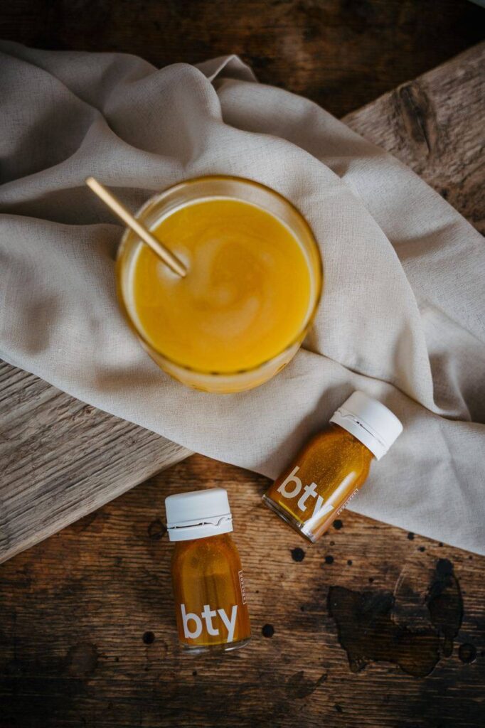 Ein Glas mit gelb-orangem RINGANA BTY (veganes Kollagen) und einem Strohhalm steht auf einem hellen Tuch, daneben zwei kleine Flaschen mit der Aufschrift bty, die veganes Kollagen enthalten, alles auf einer rustikalen Holzfläche.