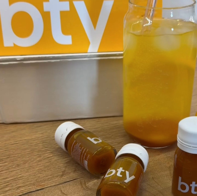 Ein Glas mit gelb-orangem RINGANA BTY (veganem Kollagen Drink), Eis und einem Strohhalm steht auf einer Holzfläche neben drei kleinen Flaschen mit der Aufschrift bty, die Kollagen vegan für Straffe Haut hervorheben. Ein gelbes und weißes bty-Schild einer Packung RINGANA BTY ist teilweise im Hintergrund zu sehen.