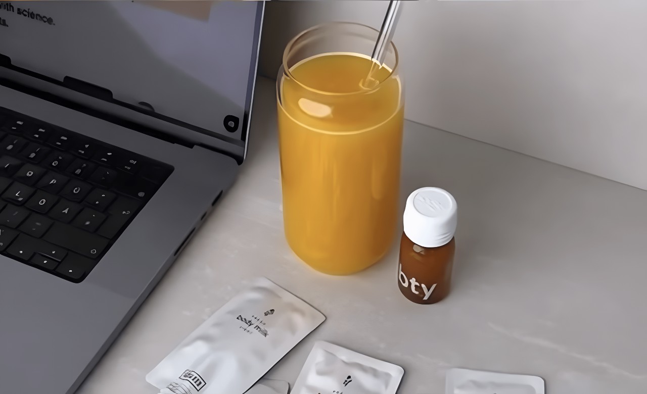 Ein Glas Orangensaft mit Strohhalm steht neben einer kleinen braunen Flasche veganes Kollagen, mehreren weißen Päckchen und einem aufgeklappten Laptop auf einem hellen Schreibtisch.
