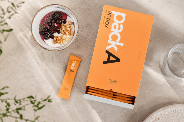 Neben einer Schale mit Joghurt, Beeren, Banane, Chiasamen und Walnüssen steht eine Schachtel mit der Aufschrift packA antiox mit orangen Einzelportionspackungen. Entdecken Sie die Frische der RINGANA Frischekosmetik Produkte, während ein Glas Wasser und eine Grünpflanze die Szene vervollständigen.